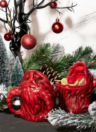 Festive scene with red ornament-laden branches, pinecones, frosted trees, and unique red mugs surrounded by greenery.