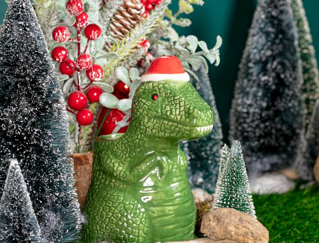 A green dinosaur figurine with a Santa hat is surrounded by snowy trees and festive greenery on a grassy surface.