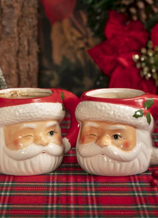 Two Santa-shaped mugs on a plaid tablecloth, with holiday decorations and a small tree in the background.