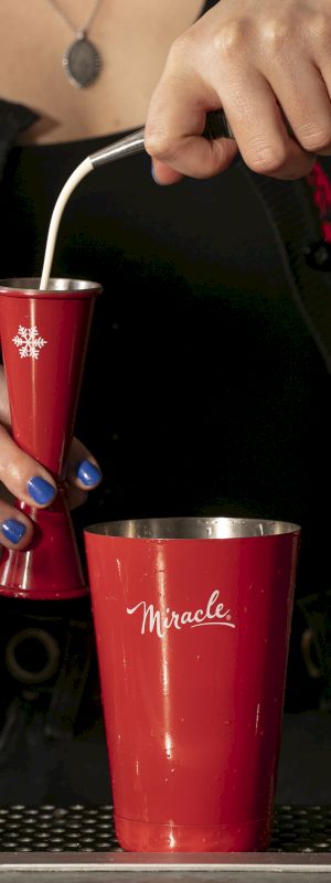 A person pouring liquid into a red measuring cup at a bar, with more red cups and decorated glasses on the counter.