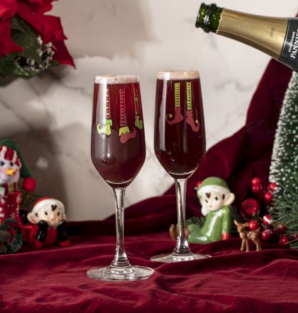 Two festive champagne glasses with holiday decorations, surrounded by Christmas-themed figurines and decor on a red cloth.