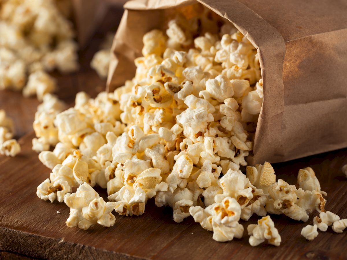 A spilled brown paper bag of freshly popped popcorn rests on a wooden surface, with kernels scattered around it.