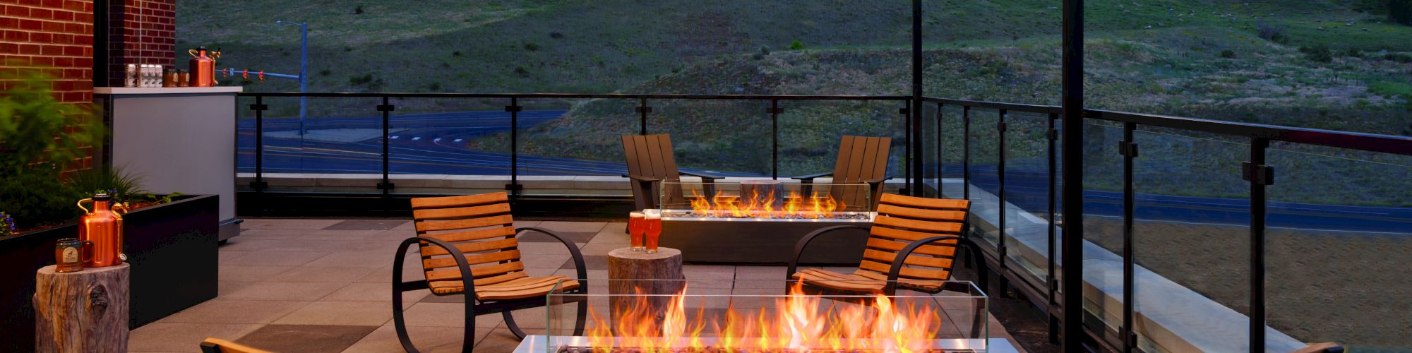 A cozy outdoor patio with several wooden chairs, fire pits, hanging string lights, and scenic hills in the background at dusk, ending the sentence.