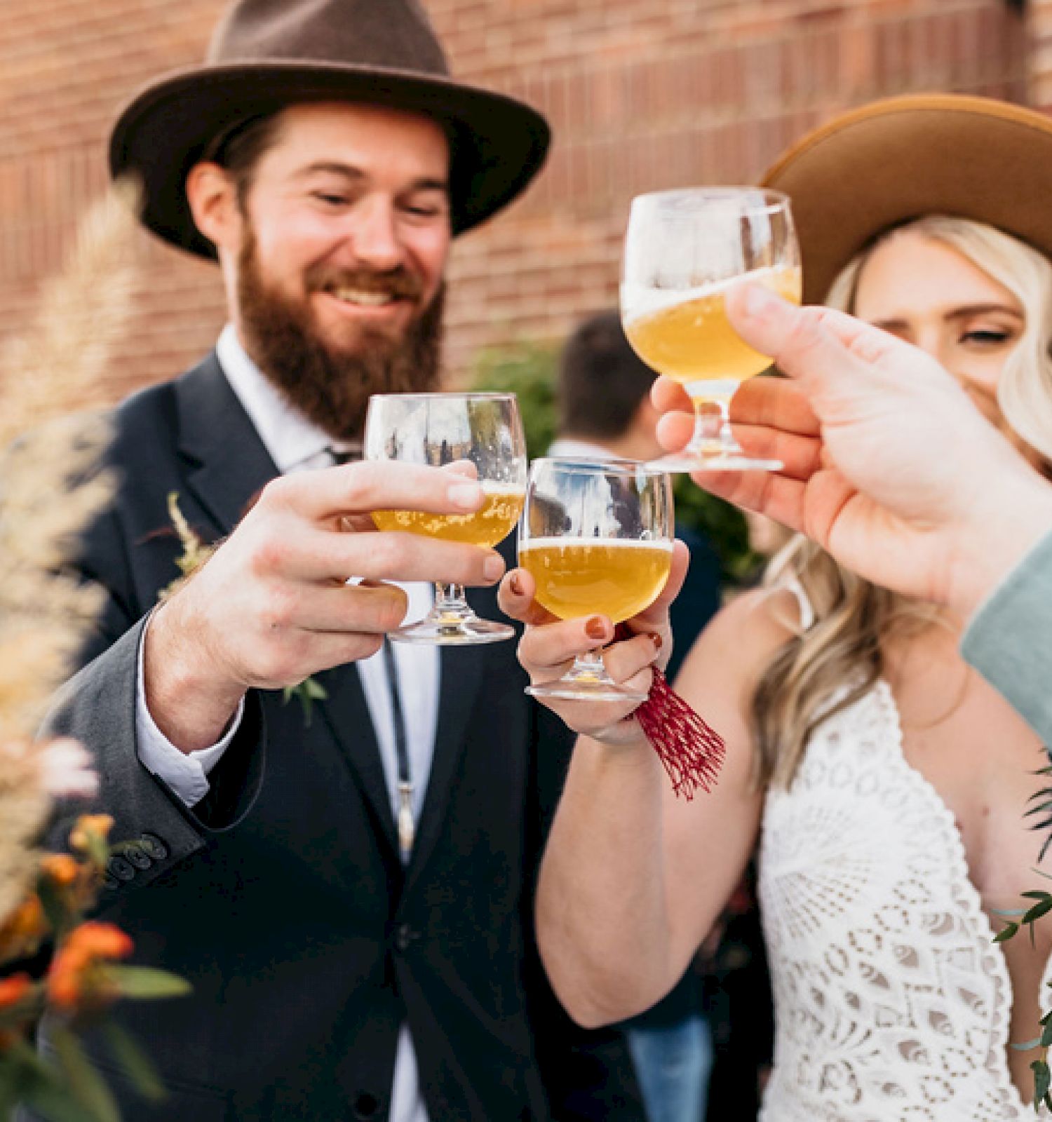 A group of people is toasting with glasses of beer or cider, dressed in formal attire and hats, near a brick wall.