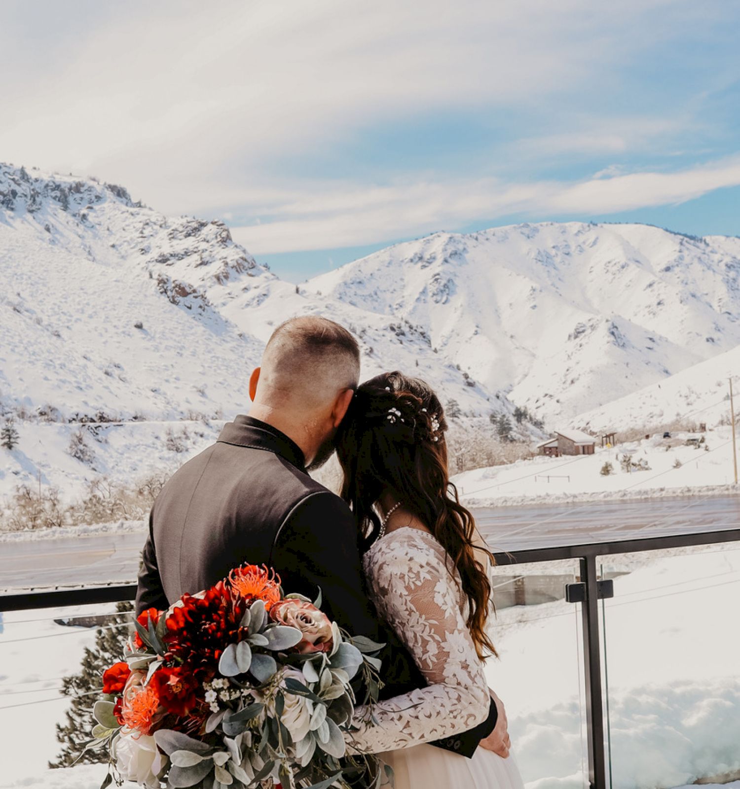 A couple embraces, the woman holding a bouquet, with a snowy mountain landscape in the background under a clear sky.