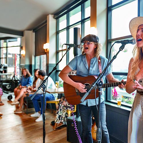 Two musicians perform in a cozy setting, with one playing guitar and the other singing. An audience sits and listens attentively.