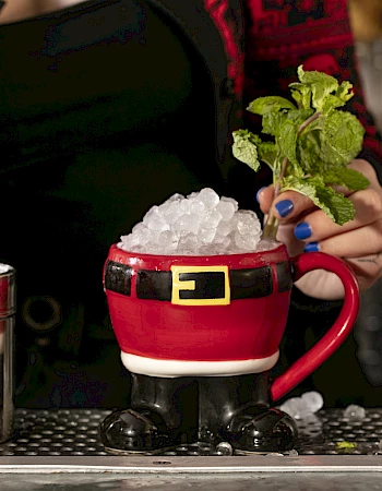 A festive cocktail in a Santa-themed mug filled with ice and mint, served by a person in a holiday sweater.