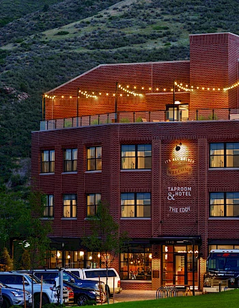 A brick hotel from the front, lights strung on the roof, a few vans parked, green hills in the background, and a welcoming entrance.